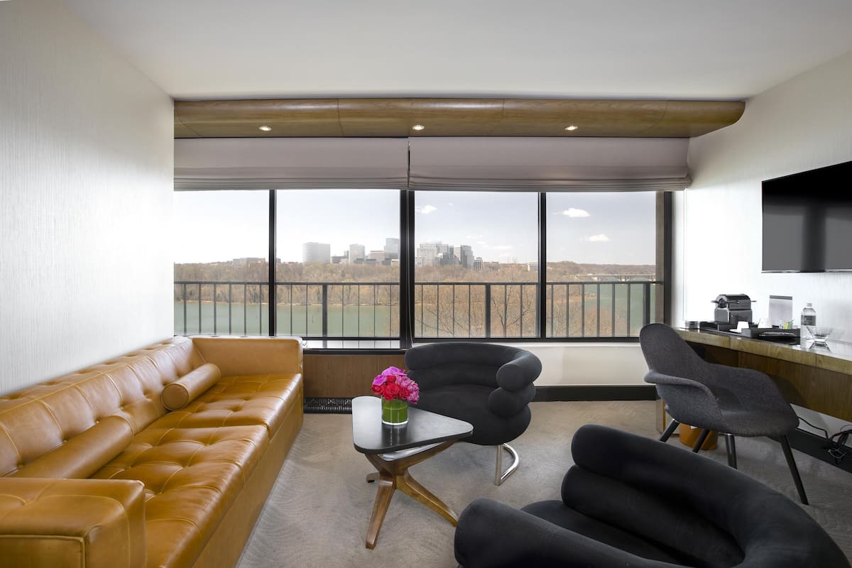 Un salon moderne avec un canapé en cuir, deux fauteuils noirs, une petite table sur laquelle est posé un vase, et une grande fenêtre donnant sur la ville.