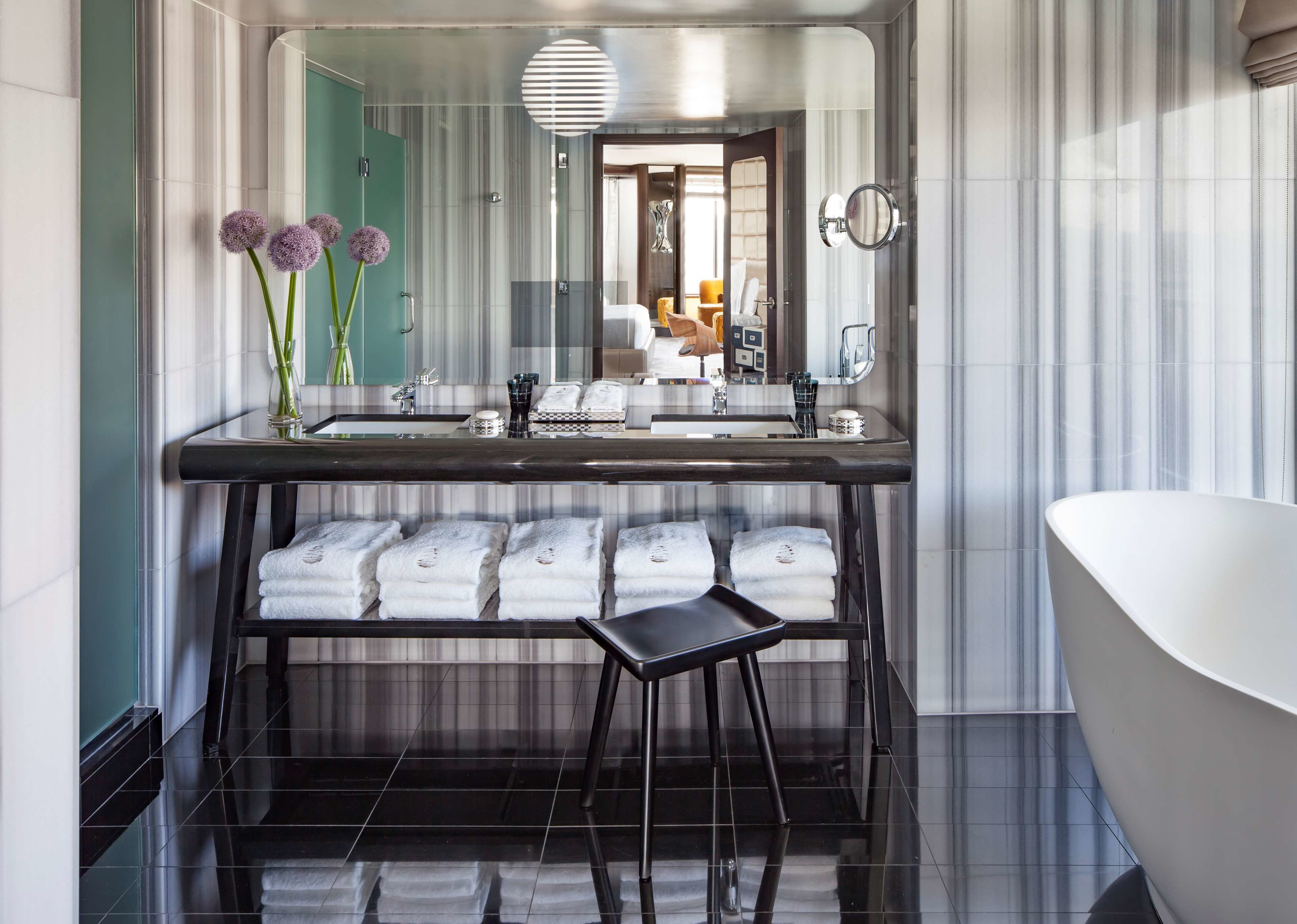 A modern bathroom with a large mirror, a bathtub, and a vanity with a sink. There are towels on a shelf and a stool in front of the vanity.