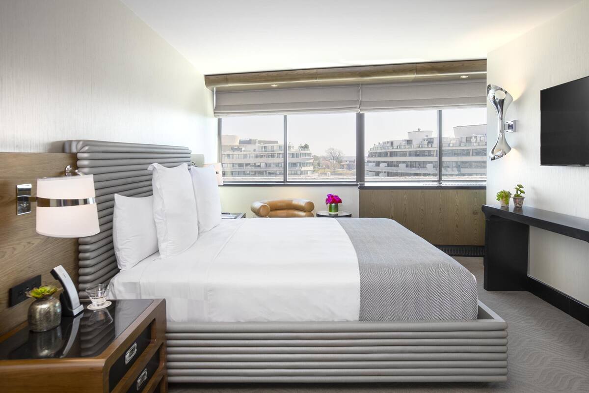 Une chambre d’hôtel moderne dotée d’un grand lit, d’une table de chevet avec une lampe, d’un téléviseur fixé au mur et d’une fenêtre donnant sur la ville.