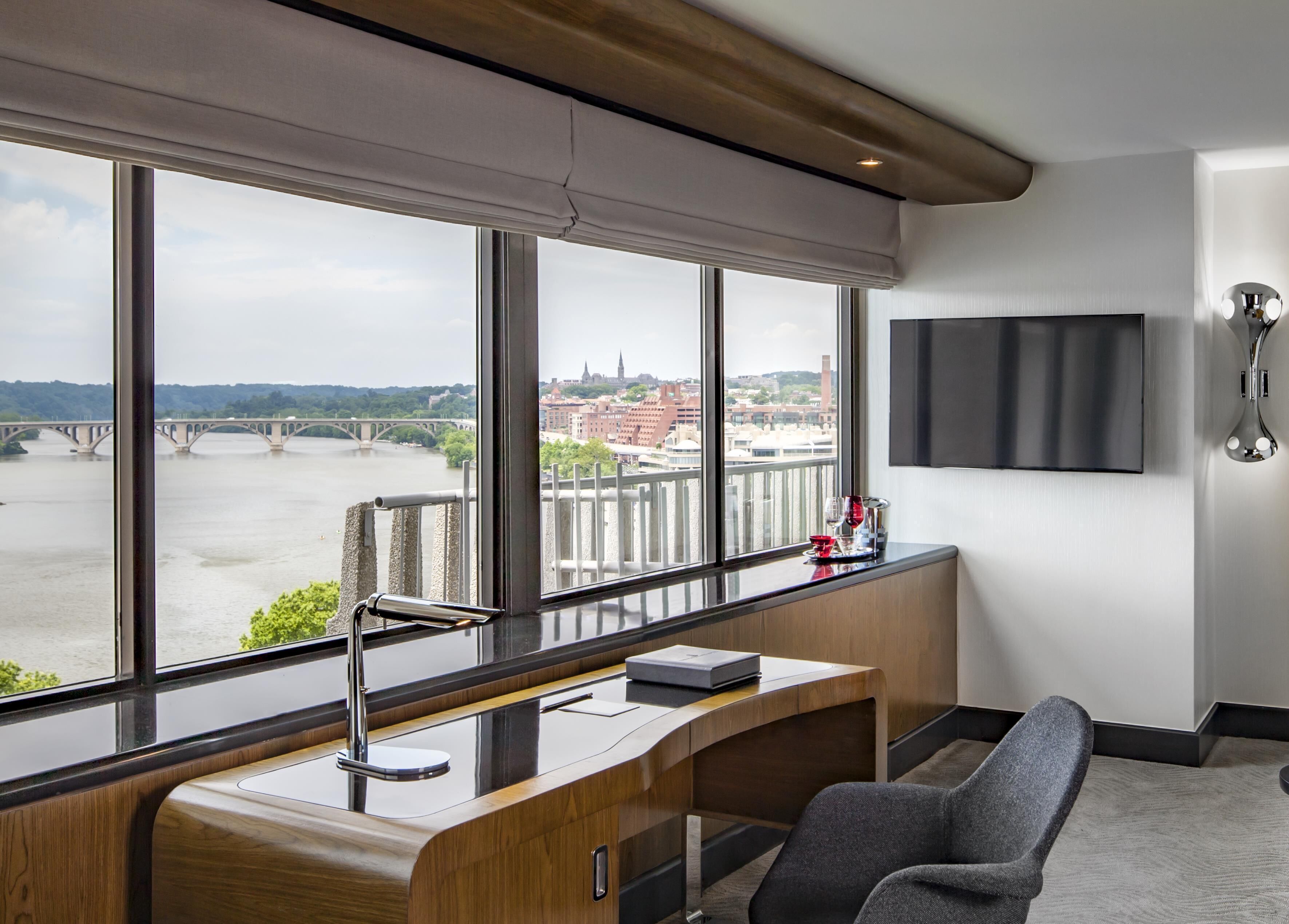 A modern office with a large window overlooking a river and a bridge, a desk with a chair, and a mounted TV.