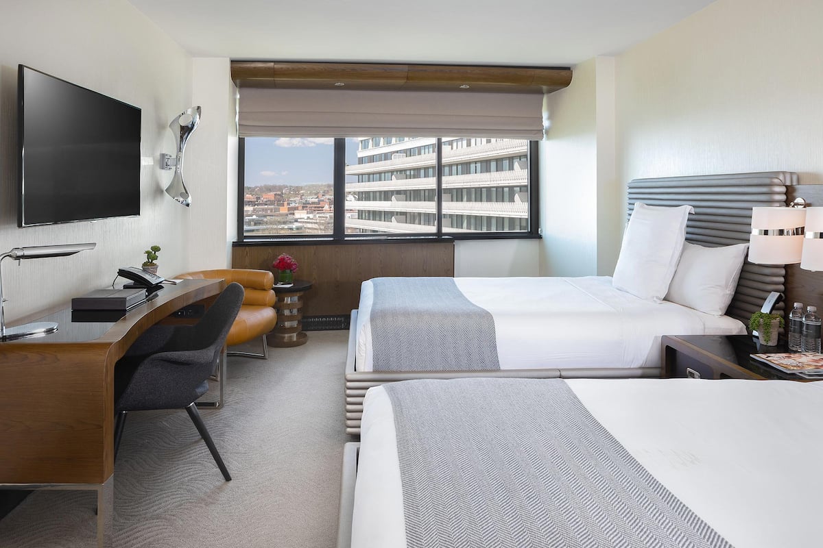 Une chambre d’hôtel équipée d’un bureau, de deux lits, d’une télévision et offrant une vue sur un immeuble.