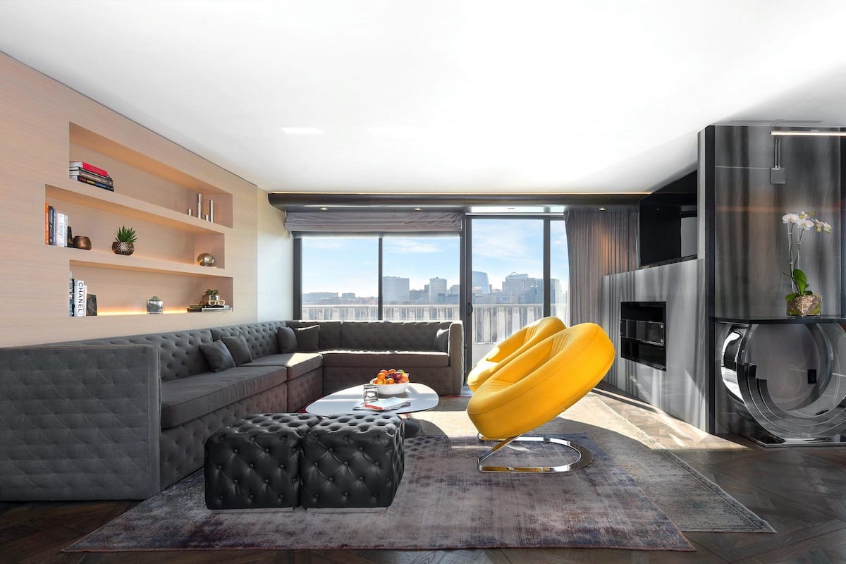 Un salon moderne avec un canapé d’angle gris, un pouf noir à capitons, un fauteuil jaune et une grande fenêtre donnant sur la ville.