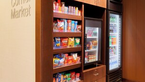 Uma mercearia de esquina com prateleiras repletas de lanches e um refrigerador com bebidas.