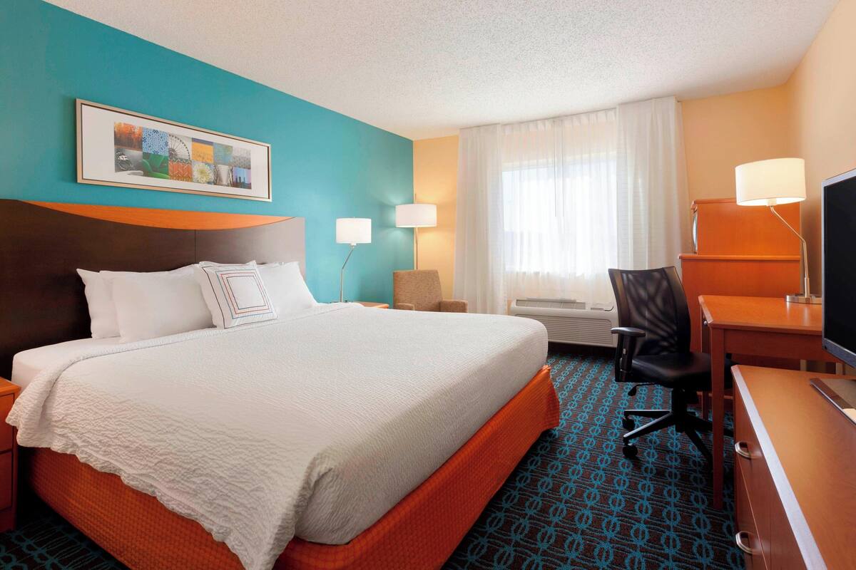 Ein Hotelzimmer mit einem großen Bett, einem Schreibtisch mit Fernseher, einem Sessel und einer Schreibtischlampe.