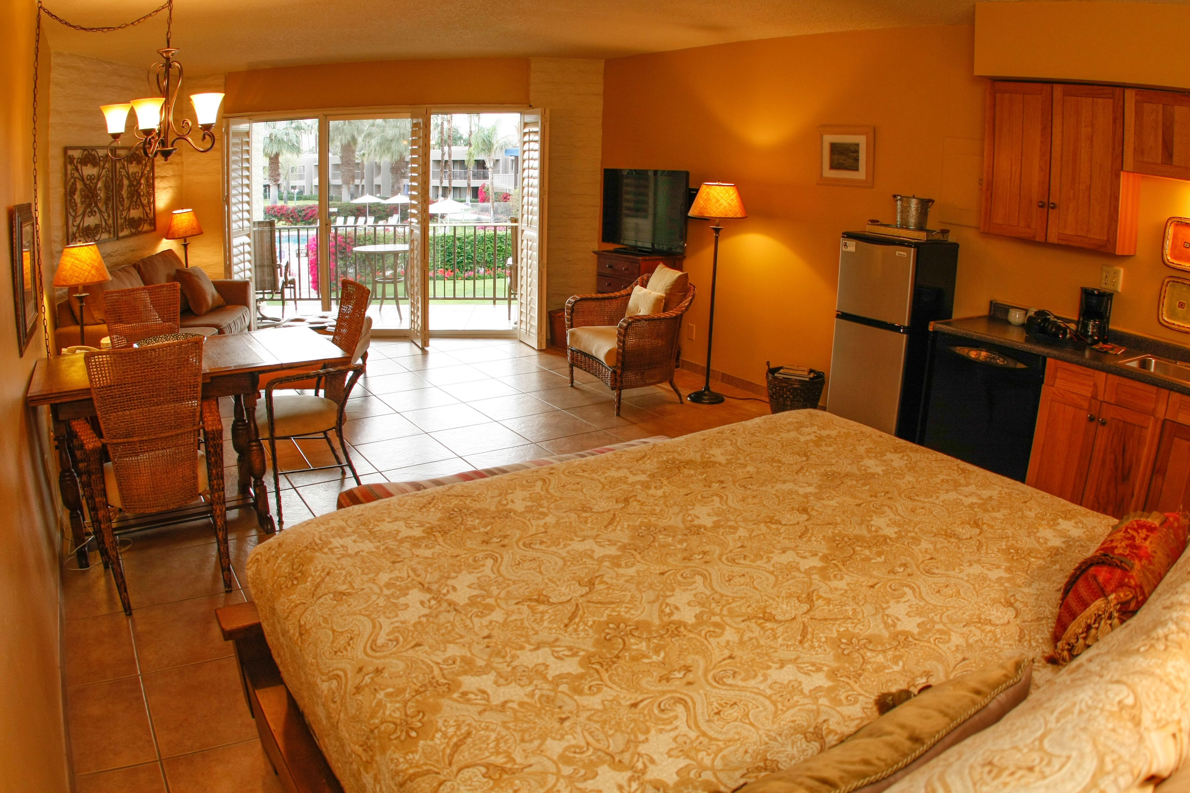 Una camera d'albergo con un letto grande, una zona pranzo con sedie e un tavolo, un angolo cottura con mobili e un frigorifero, e un balcone con vista esterna.