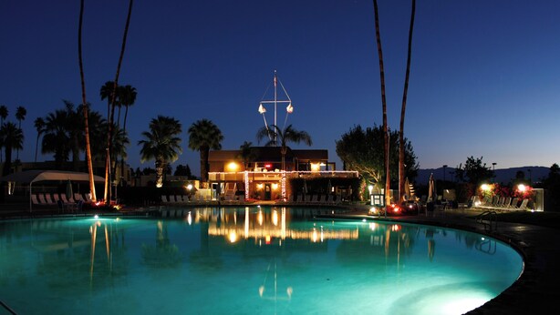 A nighttime pool area with palm trees, lounge chairs, and a building with lights. - Shadow Mountain Resort (Palm Desert)