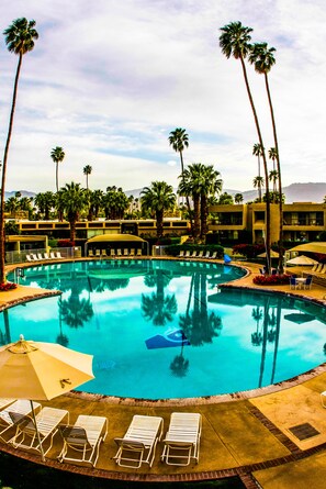 A pool with lounge chairs and umbrellas, surrounded by palm trees and a building with a curved roof. - Shadow Mountain Resort (Palm Desert)