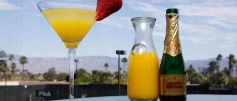 Un bicchiere da cocktail con una fragola, una bottiglia di champagne e una bottiglia di succo d'arancia su un tavolo con vista su palme e montagne.