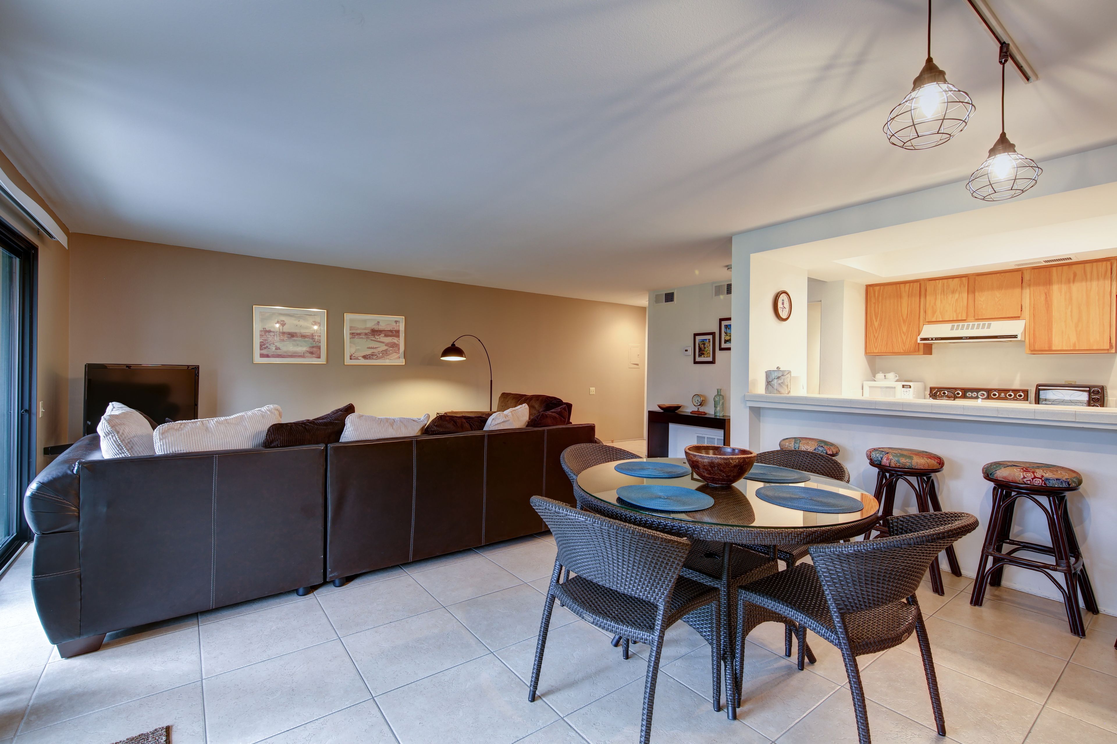 A modern living room with a dark sofa, a round dining table with wicker chairs, and a kitchen area with wooden cabinets and a built-in stove.