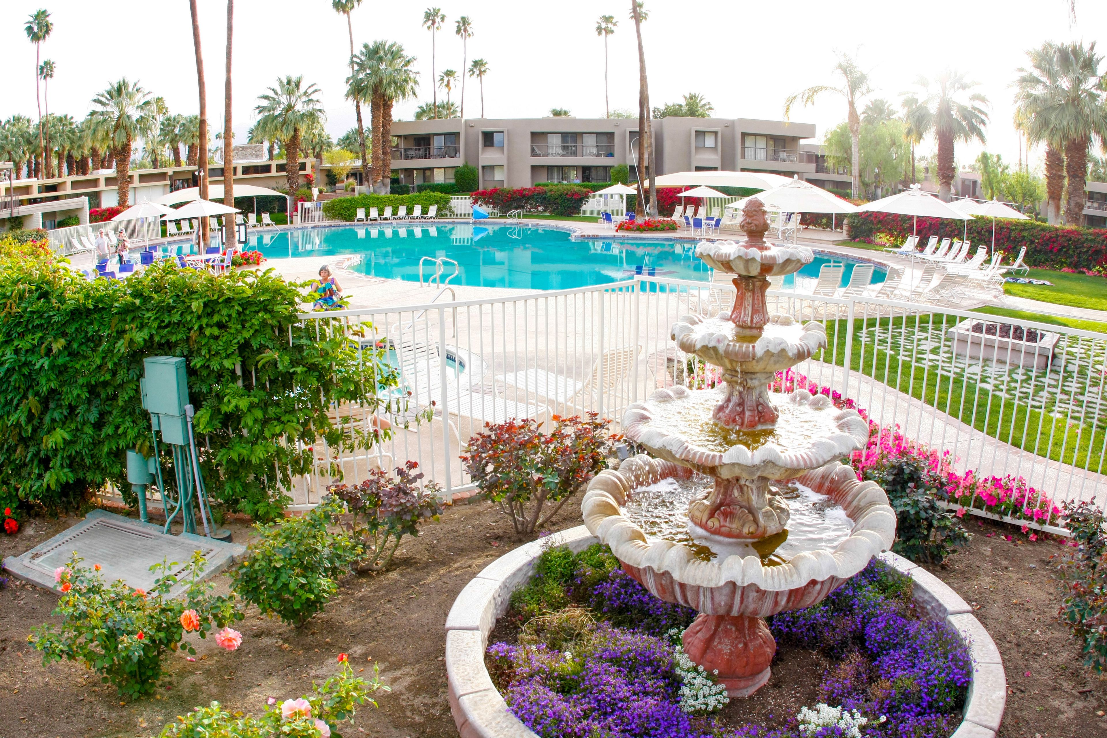 A pool area with a central fountain, surrounded by a white fence, and adjacent to a building with balconies.
