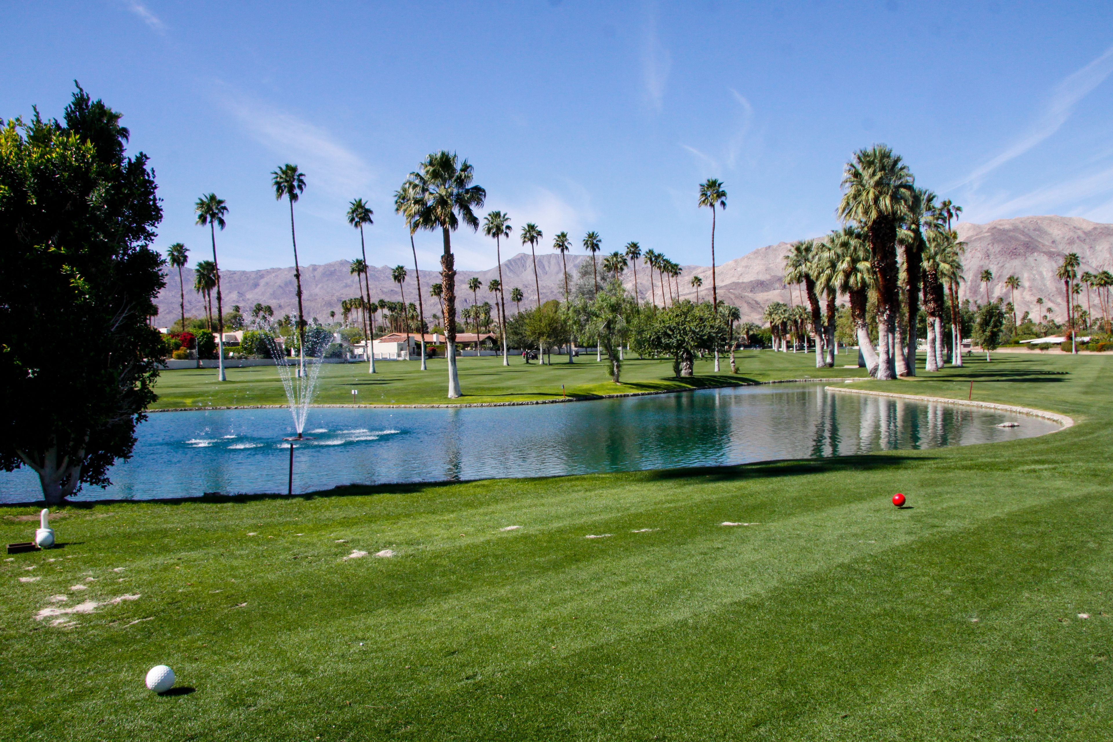 Un campo de golf con un estanque, palmeras y montañas al fondo.