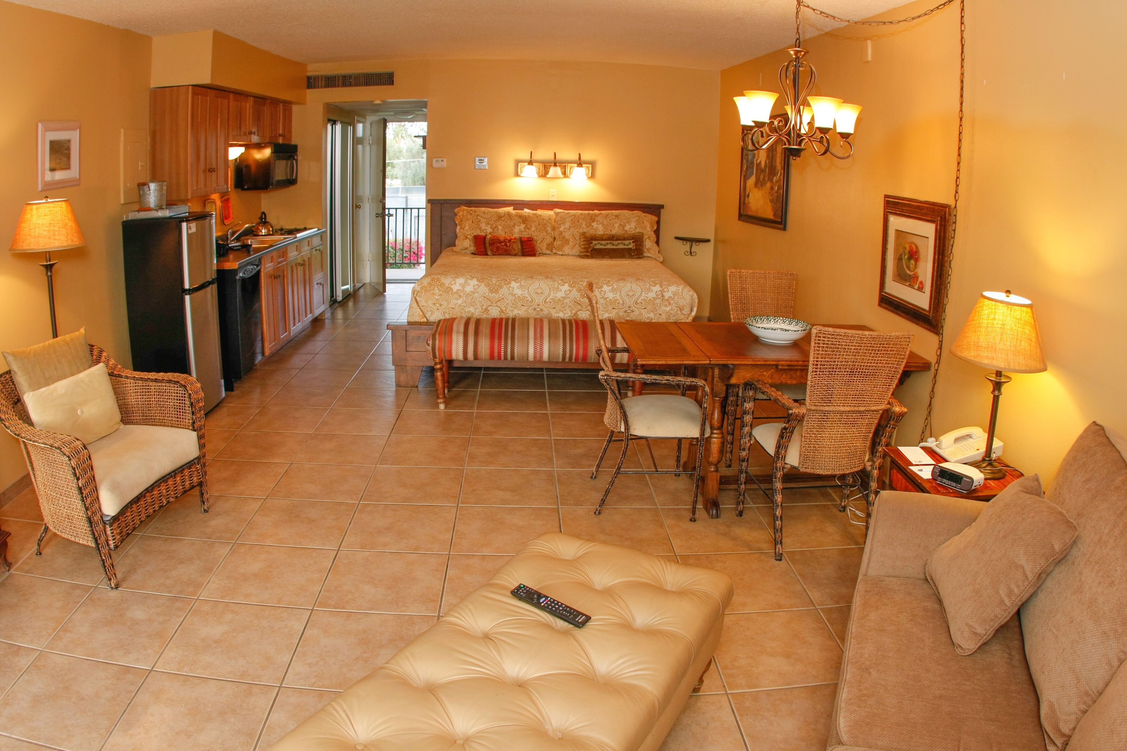 A hotel room with a bed, a dining table, a sofa, a chair, a remote control on a ottoman, and a kitchen area.