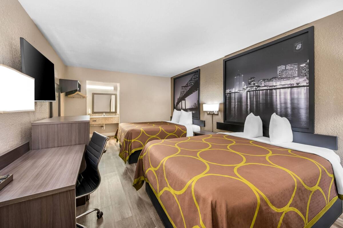 Une chambre d’hôtel avec deux lits, un bureau, une chaise, une télévision et un tableau représentant un paysage urbain.