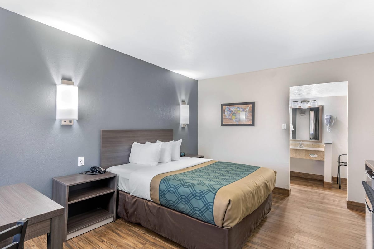 A hotel room with a bed, bedside table, lamp, and a bathroom with a sink and mirror.