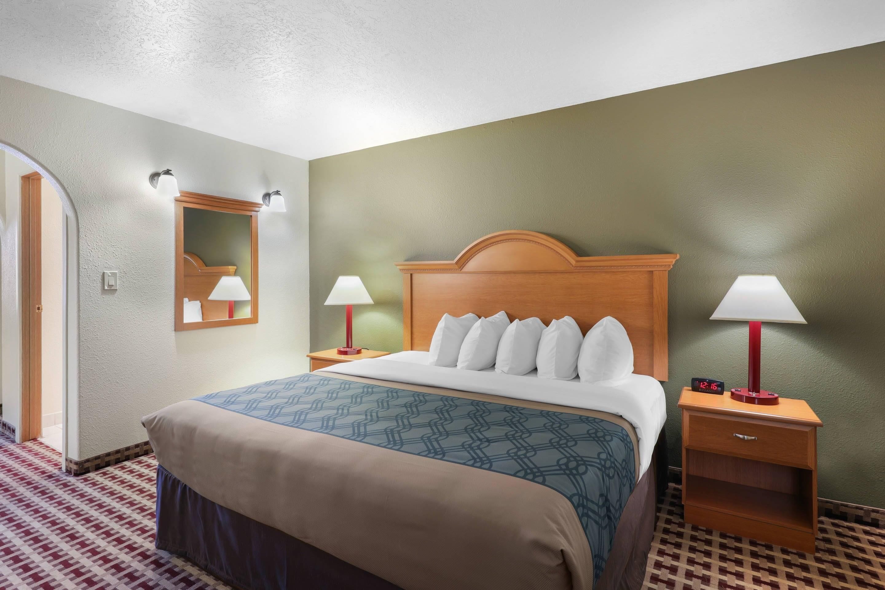 A hotel room with a bed, two bedside tables with lamps, a nightstand, a mirror, and a patterned carpet.