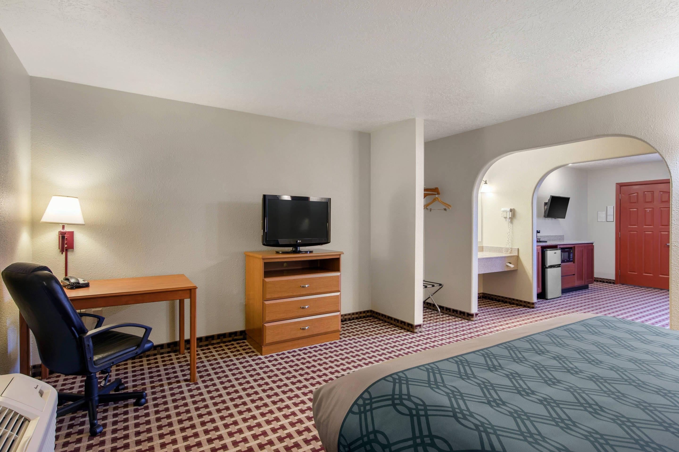 Een hotelkamer met een bed, een bureau met stoel, een televisie op een houten kast en een kitchenette op de achtergrond.