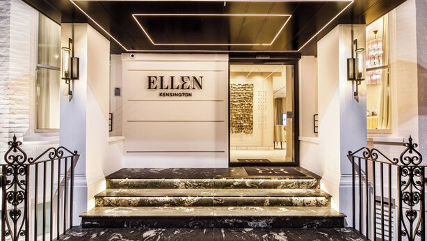 L’entrée d’un hôtel, avec un escalier en marbre, une enseigne portant le nom « ELLEN KENSINGTON » et un luminaire moderne au-dessus de l’entrée.