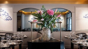 Un espace repas avec des tables rondes dressées pour le repas, un grand vase rempli de fleurs au centre, et des peintures murales représentant des nuages et des arbres.