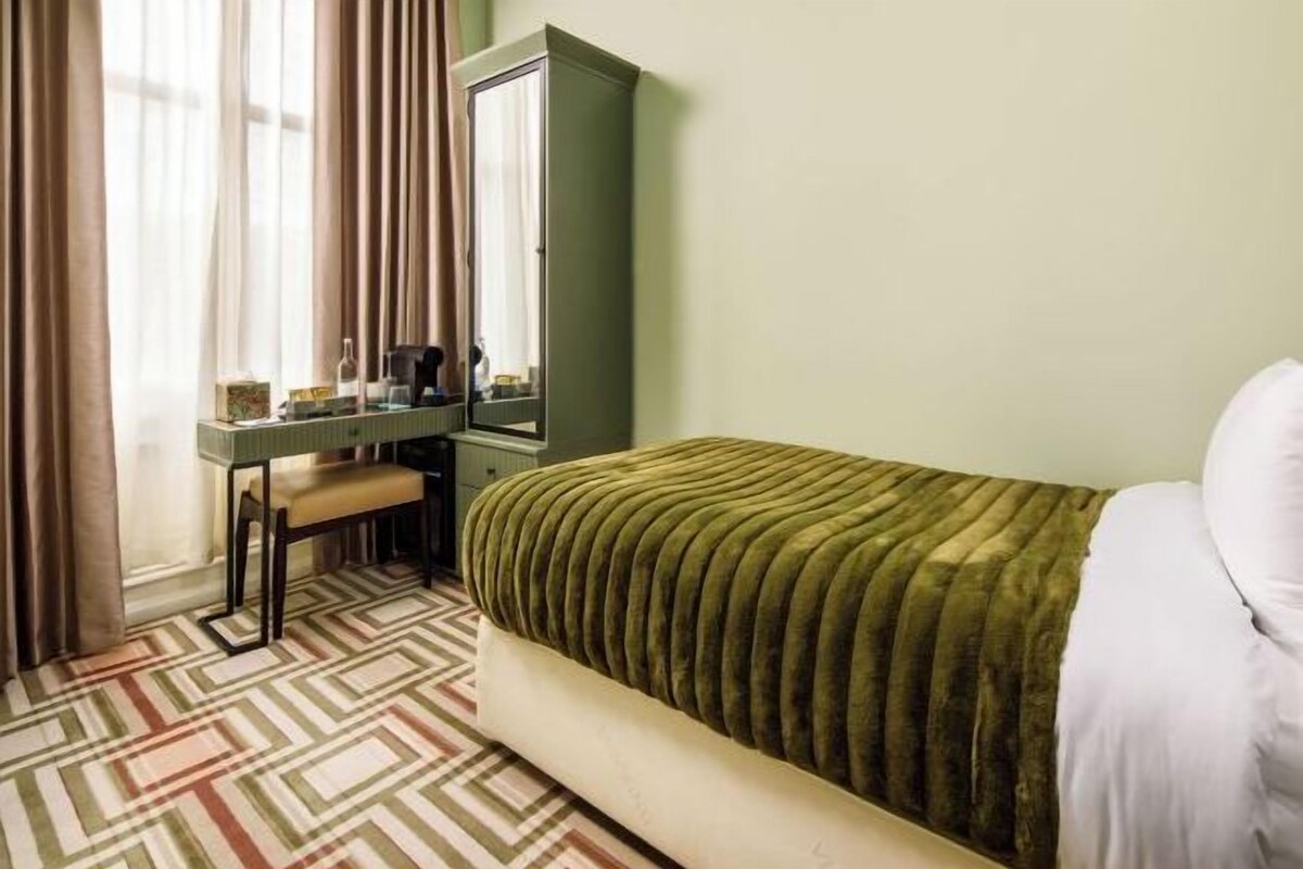 A hotel room with a bed, a desk with a chair, a mirror, and a window with curtains.