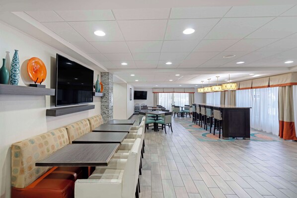 A modern restaurant with long communal tables and booths, a flat-screen TV mounted on the wall, and a patterned floor.