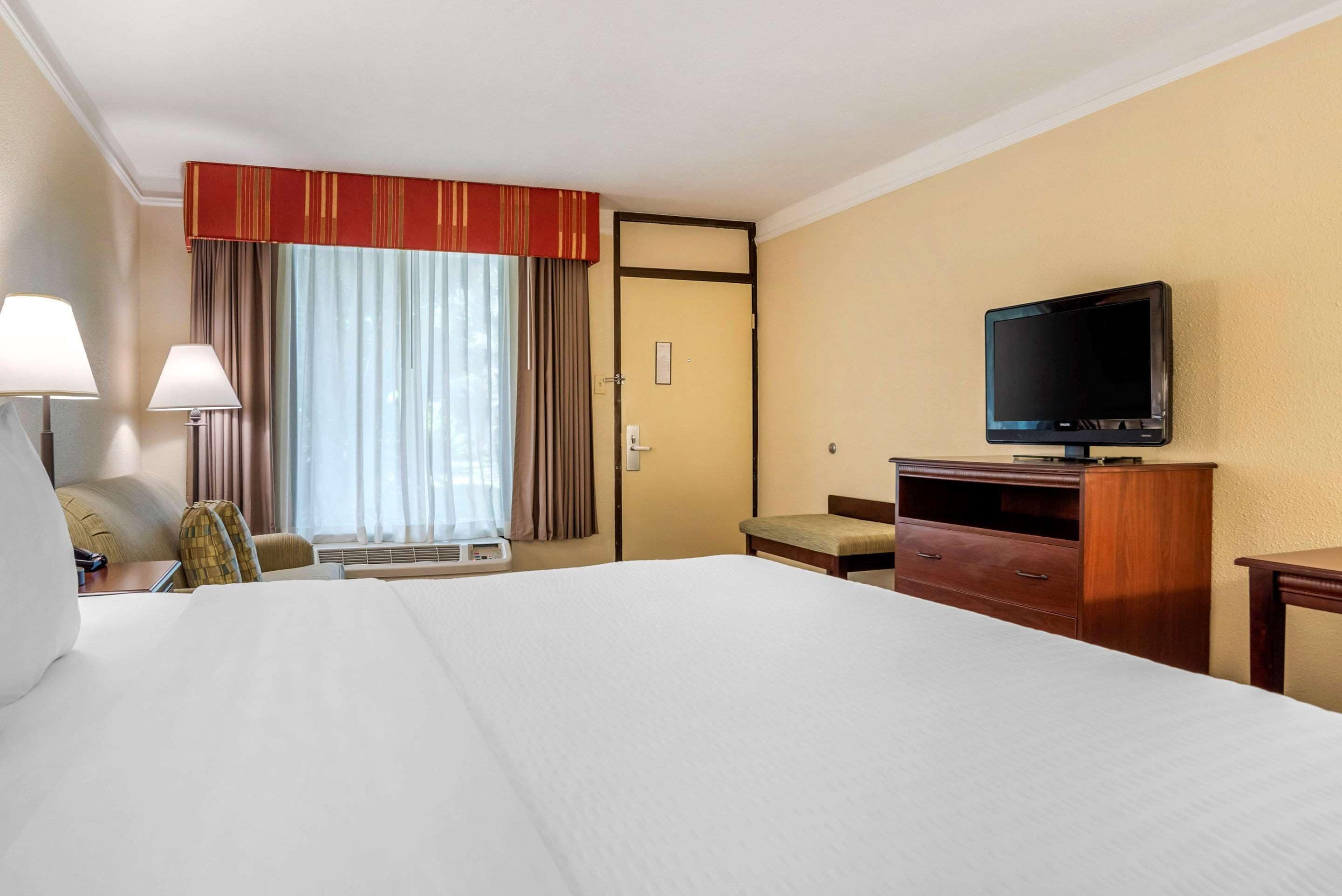 A hotel room with a large bed, a television on a wooden stand, a desk with a lamp, and a window with curtains.