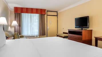 A hotel room with a large bed, a television on a wooden stand, a desk with a lamp, and a window with curtains.