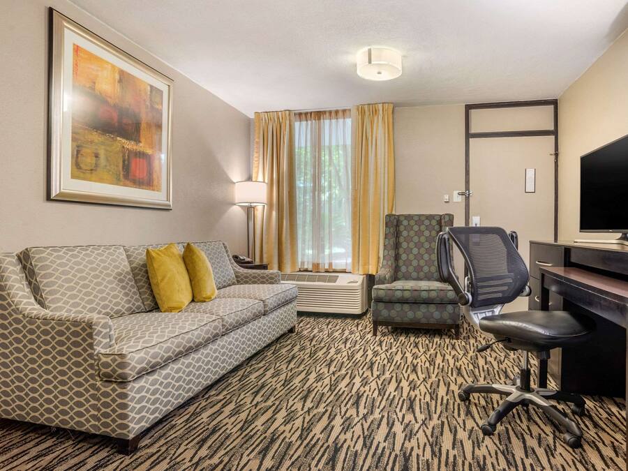 Habitación de hotel con un sofá estampado, un escritorio con silla, un televisor y un ventanal con cortinas.