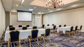 Un salón de conferencias con filas de mesas y sillas, una pantalla de proyector y una araña.