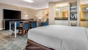 Hotellihuone, jossa on suuri sänky, työpöytä ja kaksi tuolia, taulutelevisio, minibaari sekä kylpyhuone, jossa on lasiseinällä varustettu suihkutila.