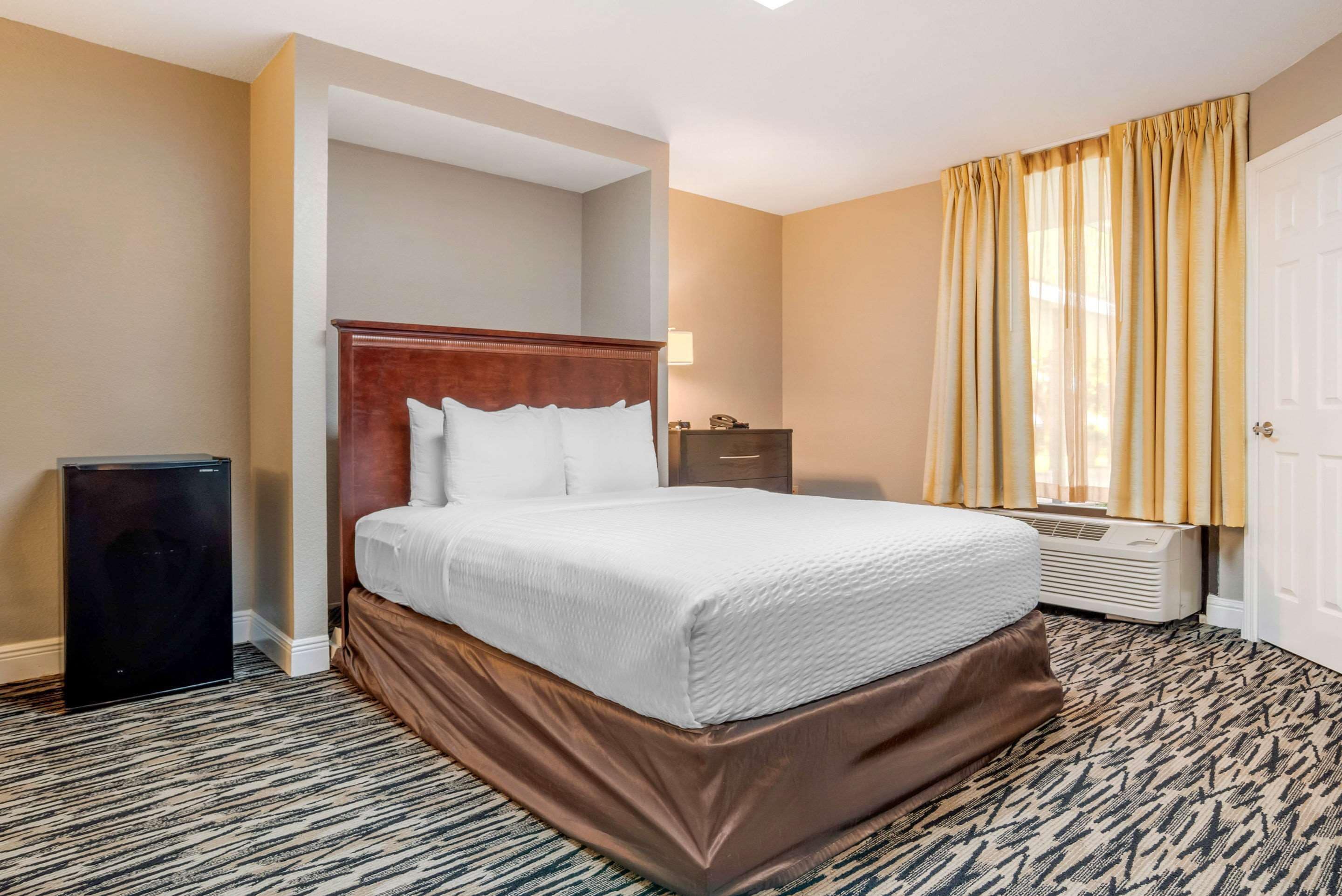 Ein Hotelzimmer mit einem großen Bett, einem Nachttisch, einer Lampe, einem Heizkörper und einem Fenster mit Vorhängen.