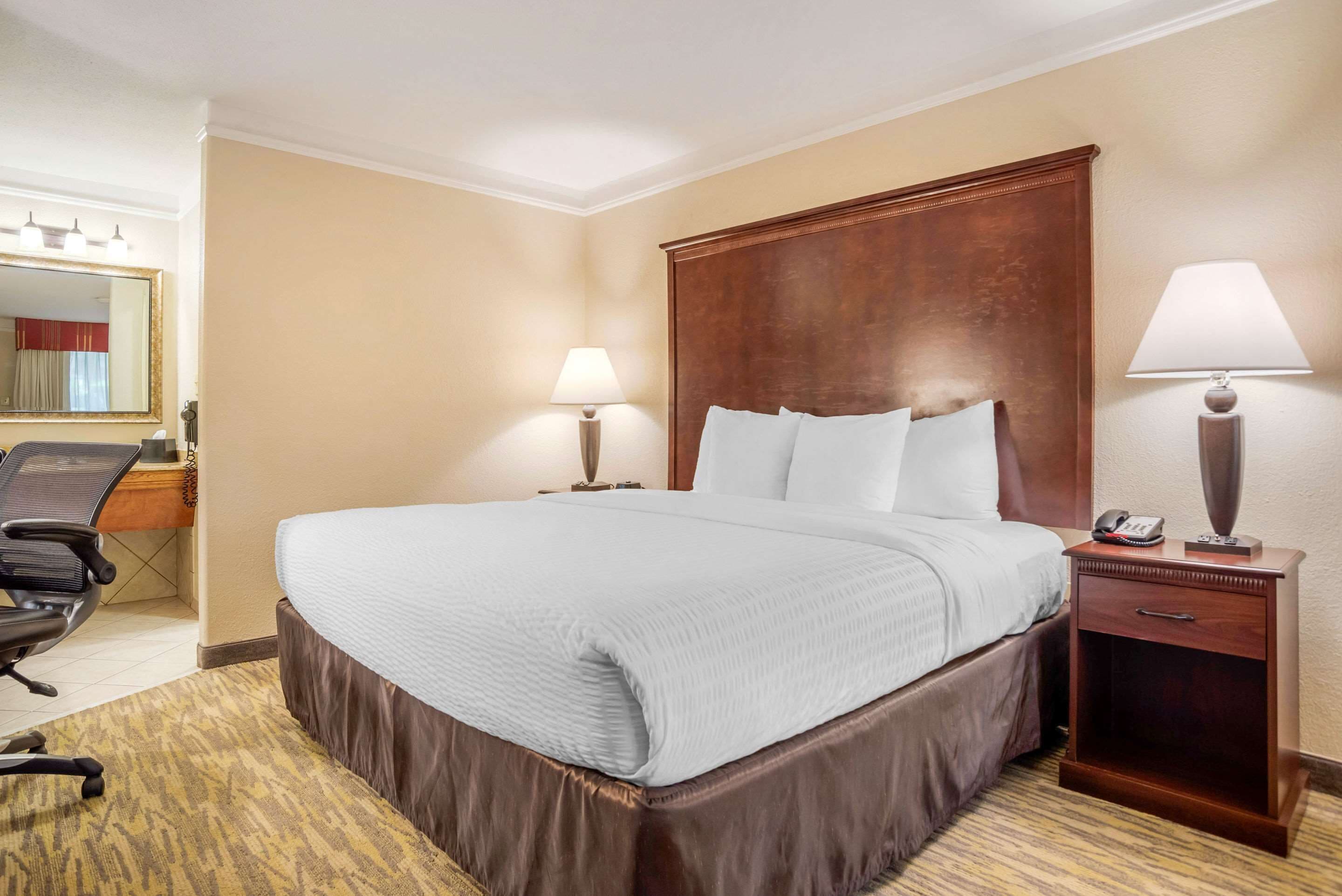 A hotel room with a large bed, two bedside lamps, a wooden nightstand, a desk with a chair, and a mirror.