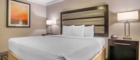 Habitación de hotel con una cama grande, una mesita de noche con lámpara, un cuadro enmarcado en la pared y un cabecero con varios almohadones.