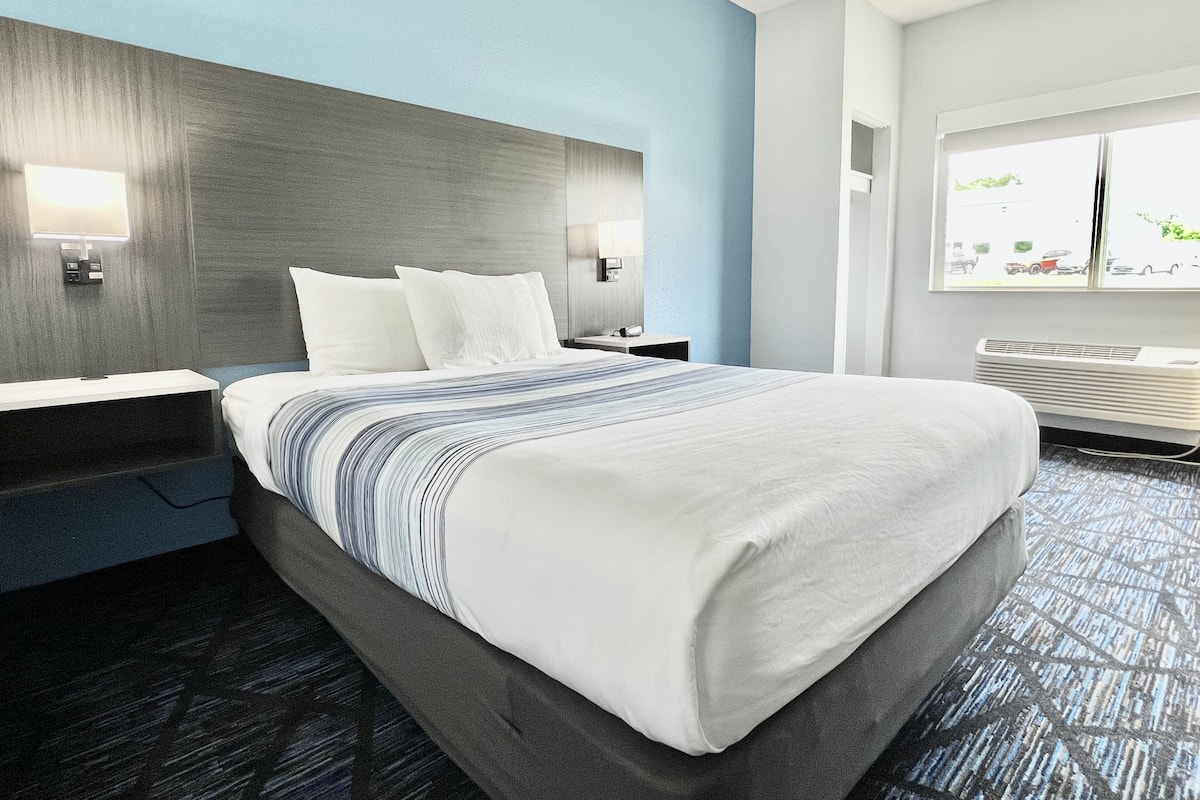A hotel room with a bed, bedside tables, a nightstand, a window with a view of the street, and a wall-mounted air conditioning unit.