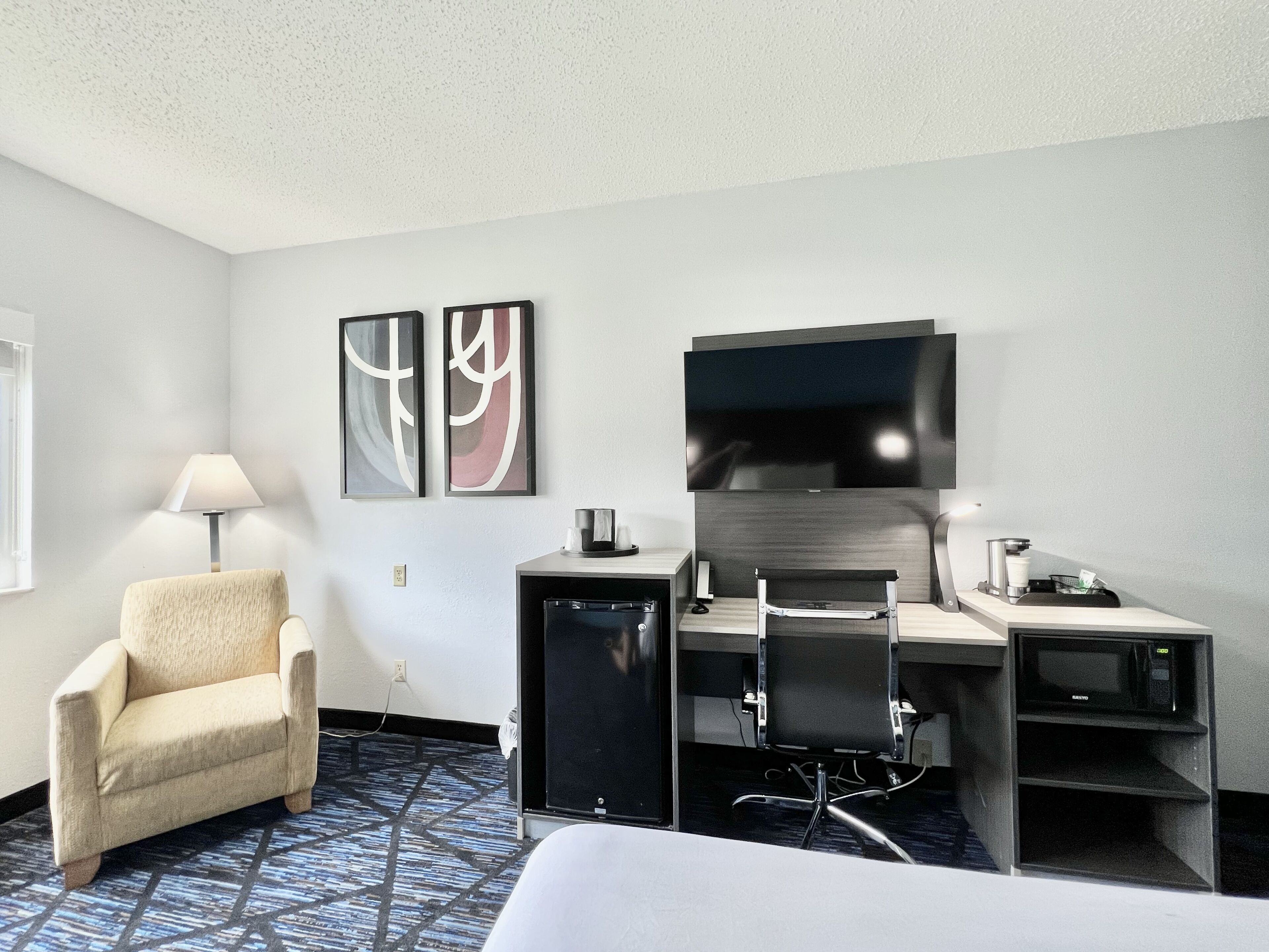 A hotel room with a bed, a desk with a chair, a TV, a lamp, and abstract wall art.