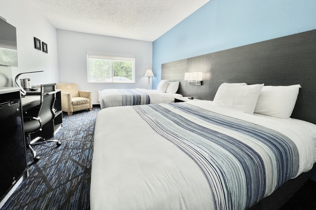 A hotel room with a large bed, a desk with a chair, a lamp, and a window with a view of greenery.