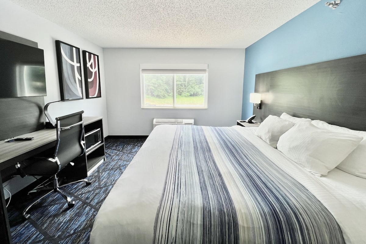 A hotel room with a bed, desk, chair, and a window with a view of greenery.