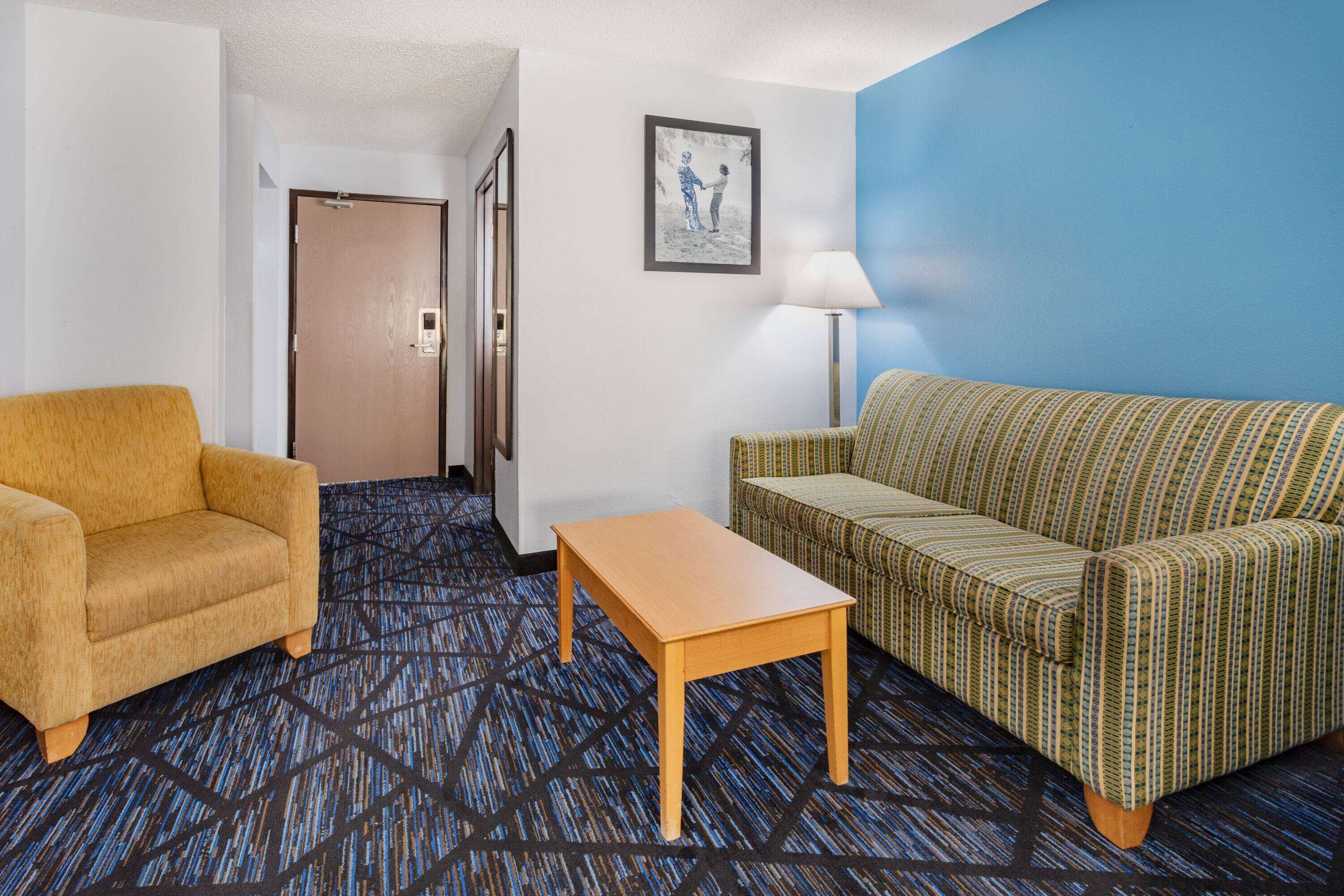 Quarto de hotel com carpete estampado, uma poltrona amarela, um sofá verde, uma mesa de centro de madeira, um quadro na parede e uma porta que dá para outro cômodo.