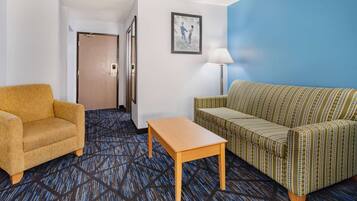 Quarto de hotel com carpete estampado, uma poltrona amarela, um sofá verde, uma mesa de centro de madeira, um quadro na parede e uma porta que dá para outro cômodo.