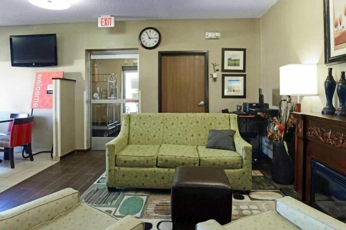 A hotel lobby with a green patterned sofa, a brown ottoman, a television, a clock, and a fireplace.