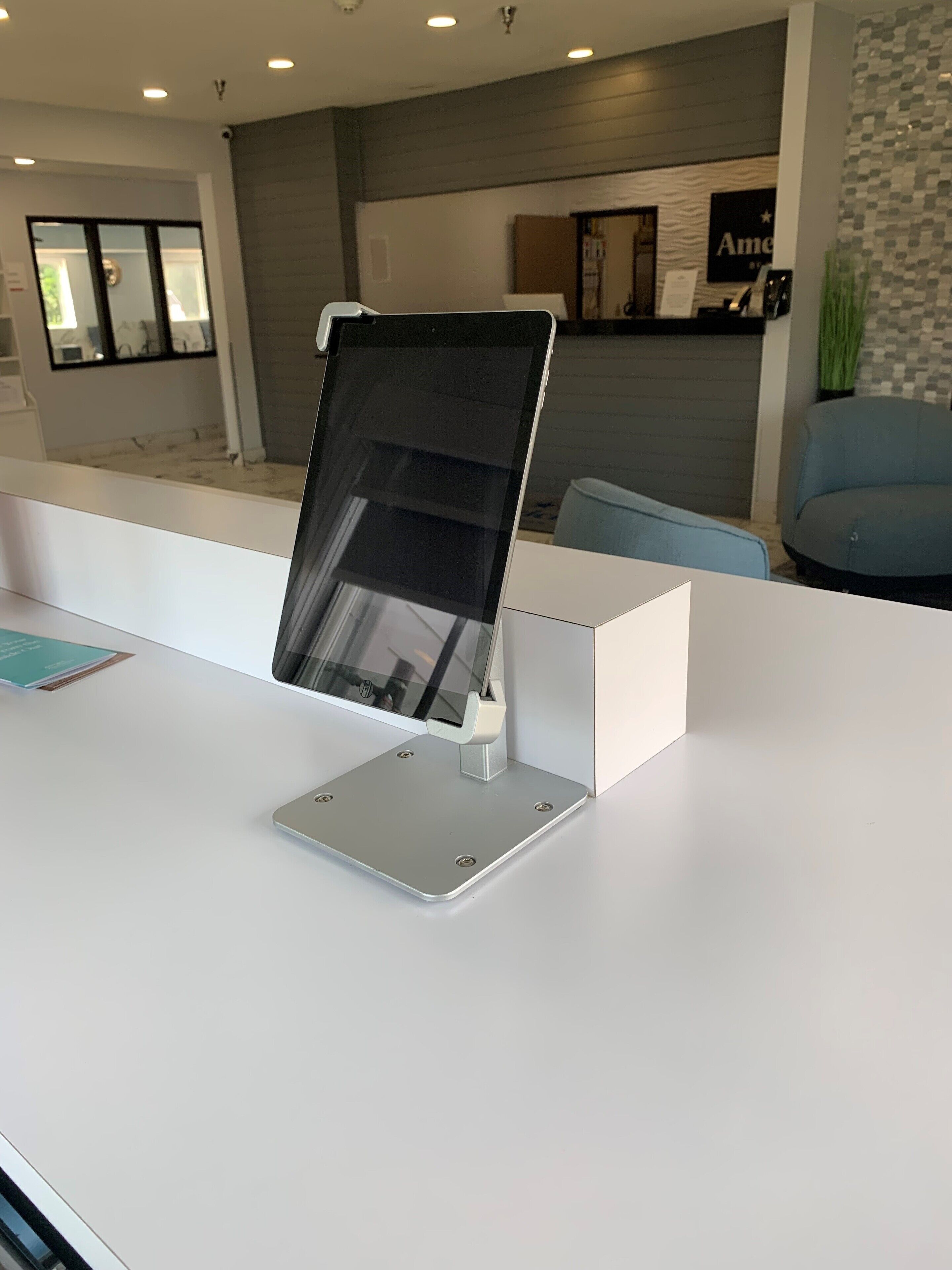 Een receptiebalie met een tablet op een standaard, een blauwe stoel, en een receptiegedeelte met een balie en een bord aan de muur.