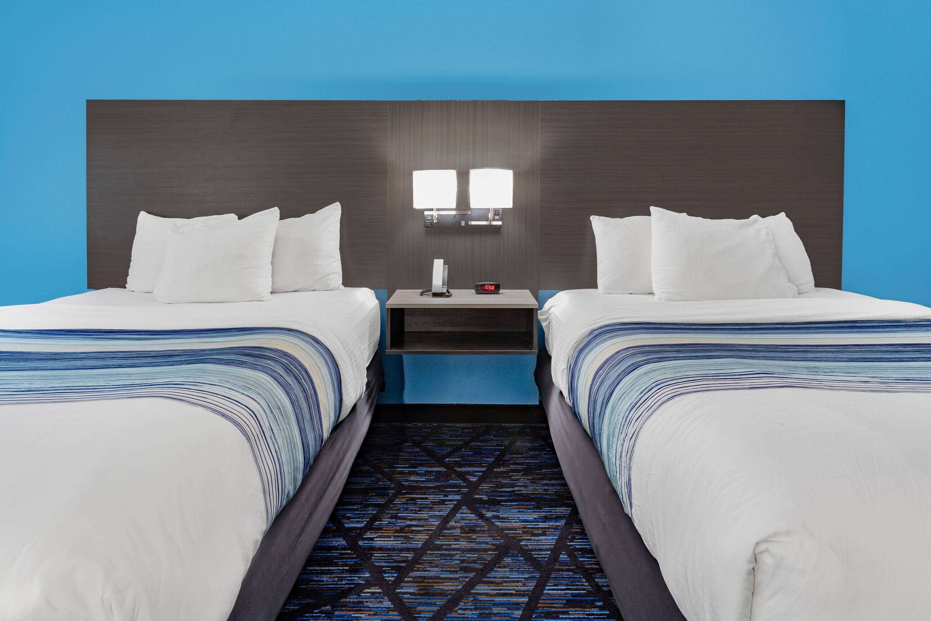 A hotel room with two beds, a nightstand with a lamp, and a blue wall.