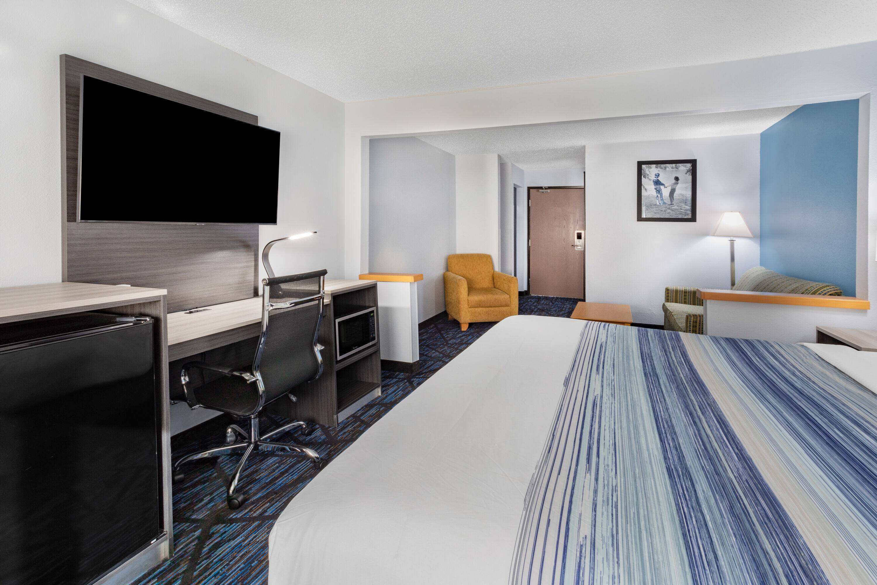Ein Hotelzimmer mit einem großen Bett, einem Schreibtisch mit Flachbildfernseher, einem Sessel, einem Nachttisch und einer Lampe.