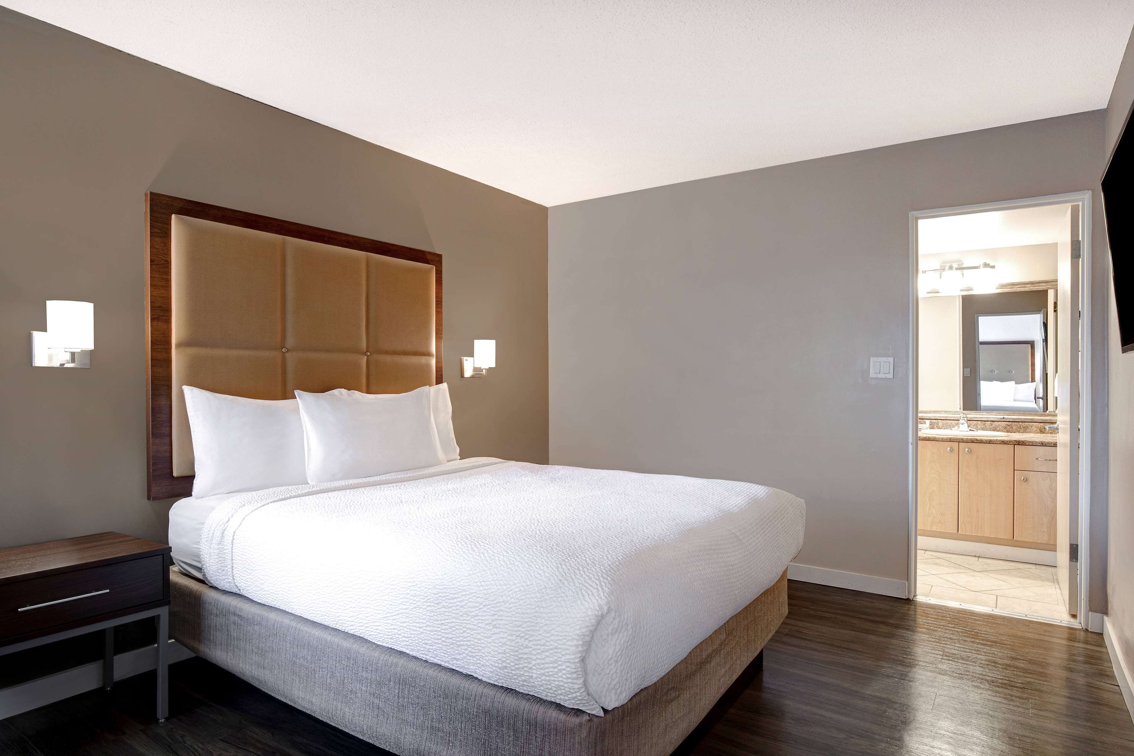 Una habitación de hotel moderna con una cama grande, un cabecero de madera, una mesita de noche y un baño visible a través de una puerta abierta.