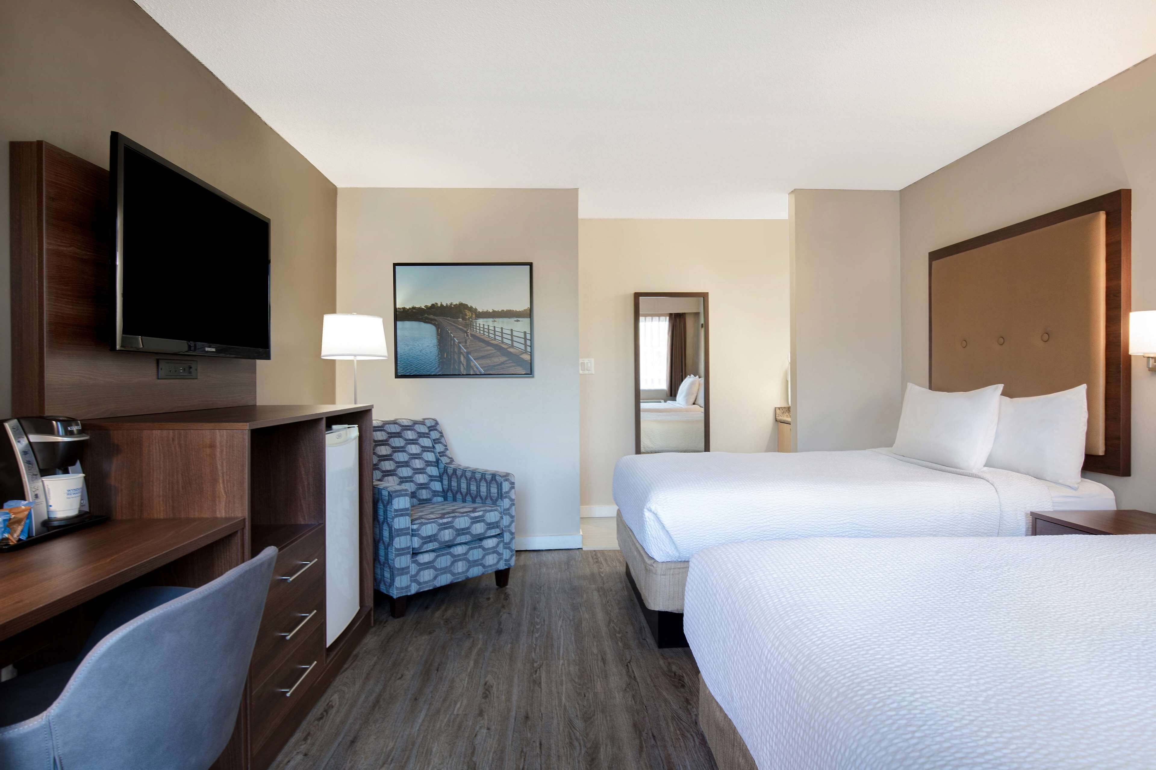 A hotel room with a flat-screen TV, a desk with a coffee maker, a patterned armchair, two beds with white bedding, and a painting of a bridge.