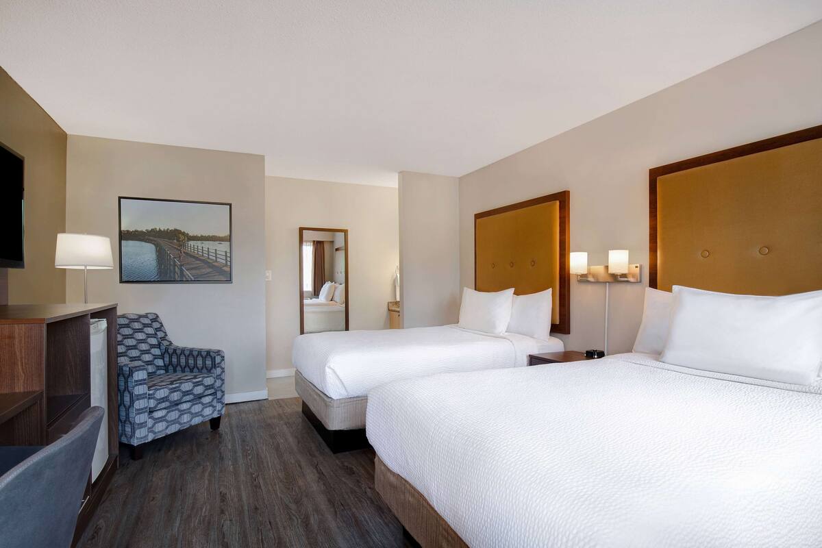 Ein Hotelzimmer mit zwei Betten, einem Schreibtisch, einem Stuhl, einem großen Spiegel und einem Gemälde eines Piers.