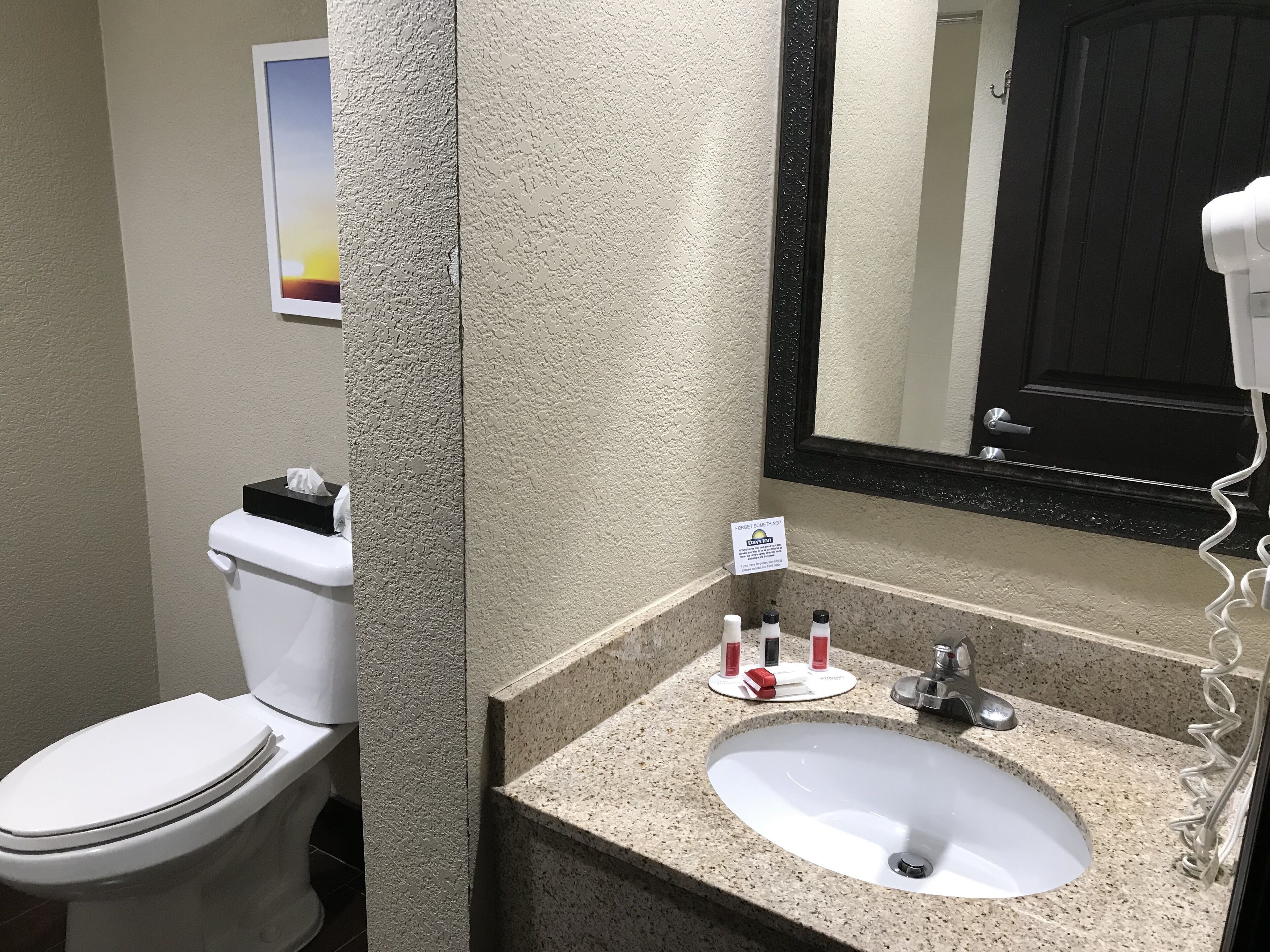 A bathroom with a toilet, sink, and mirror. There is a sign on the wall and toiletries on the sink.