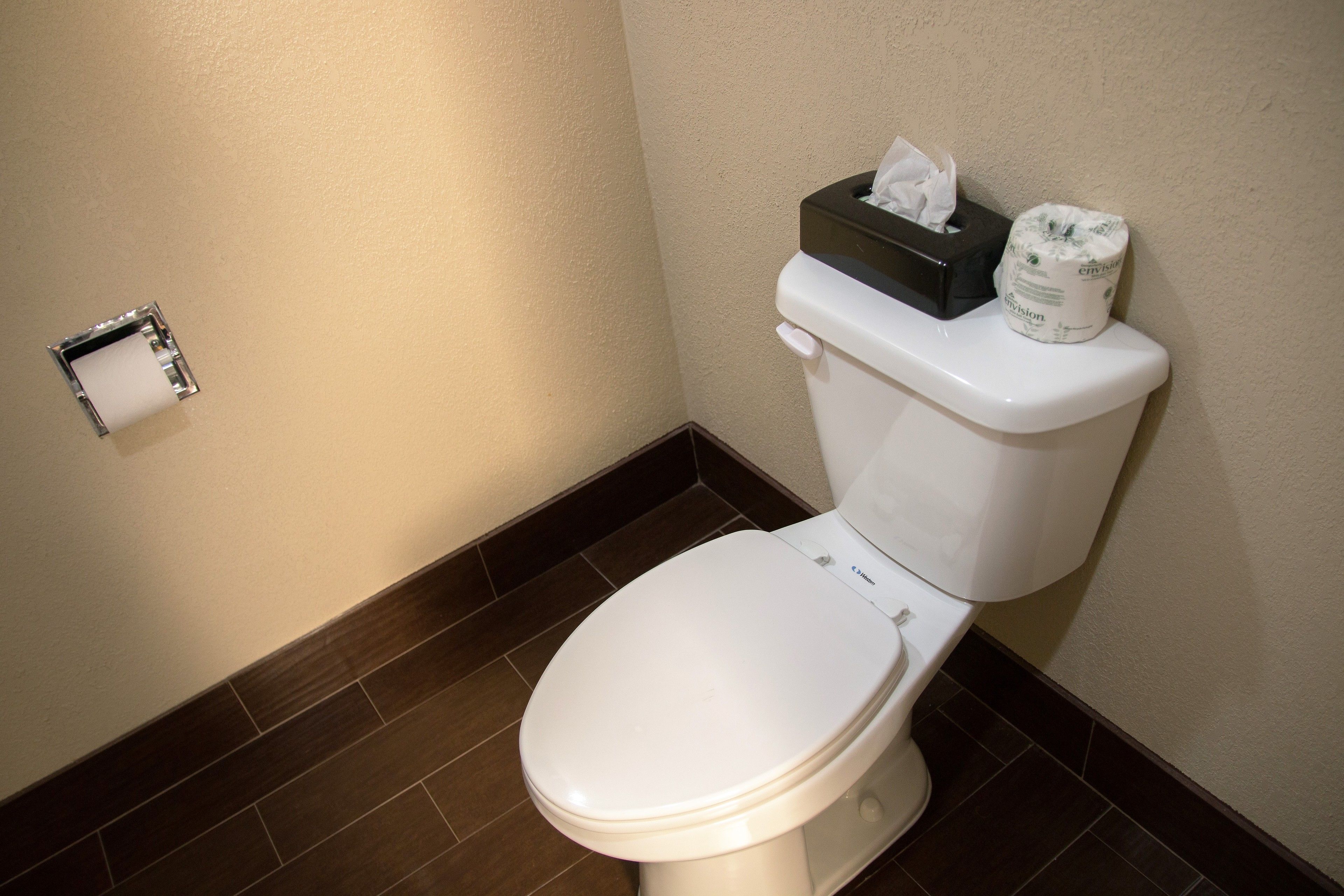 A white toilet with a tissue box and toilet paper on the tank, a toilet paper holder on the wall, and dark brown tiled flooring.