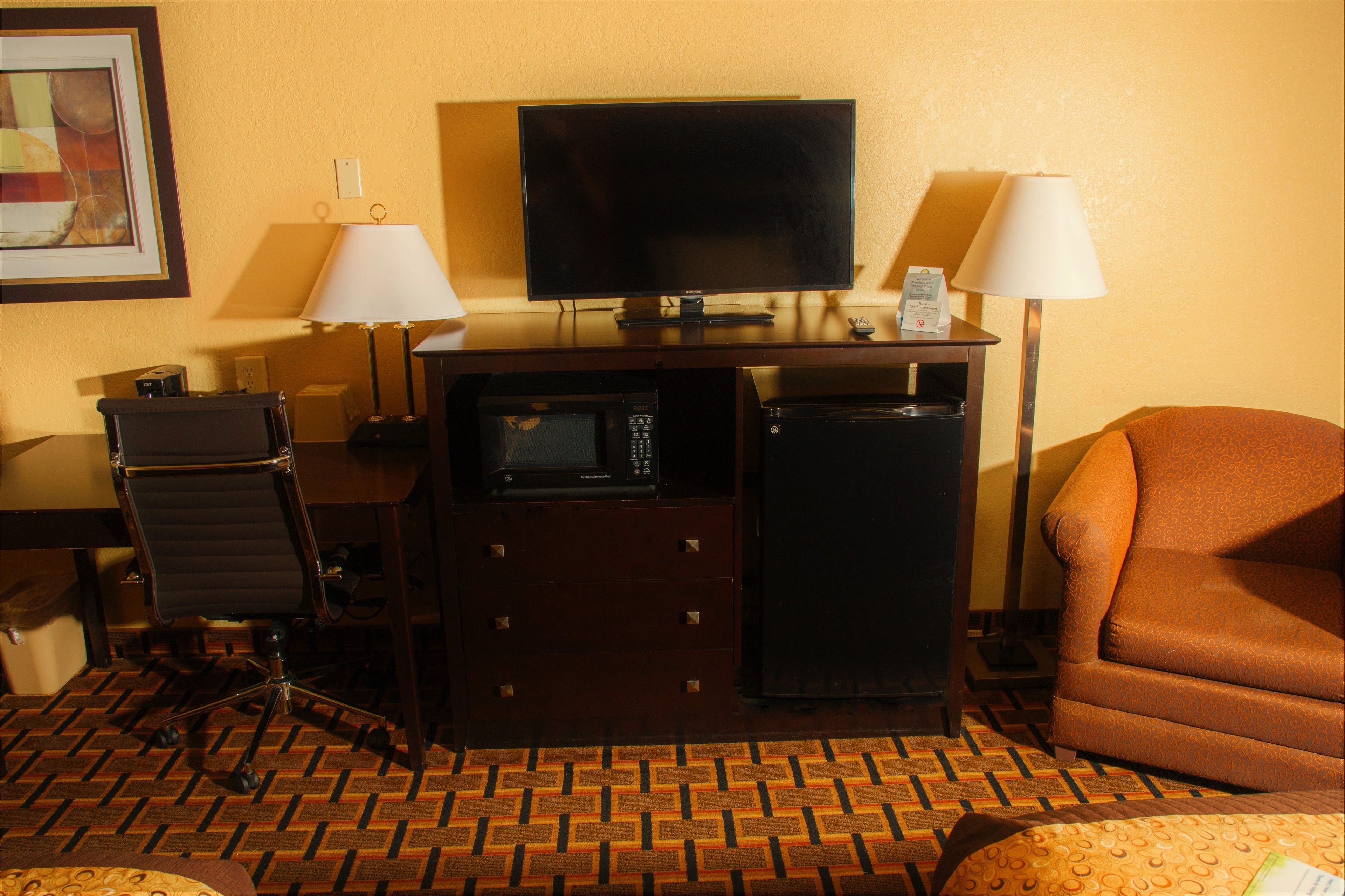 A hotel room with a television, a desk with a lamp, a microwave, a refrigerator, a chair, and a patterned carpet.