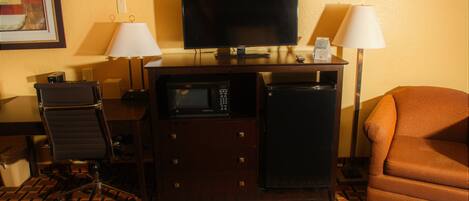 A hotel room with a television, a desk with a lamp, a microwave, a refrigerator, a chair, and a patterned carpet.