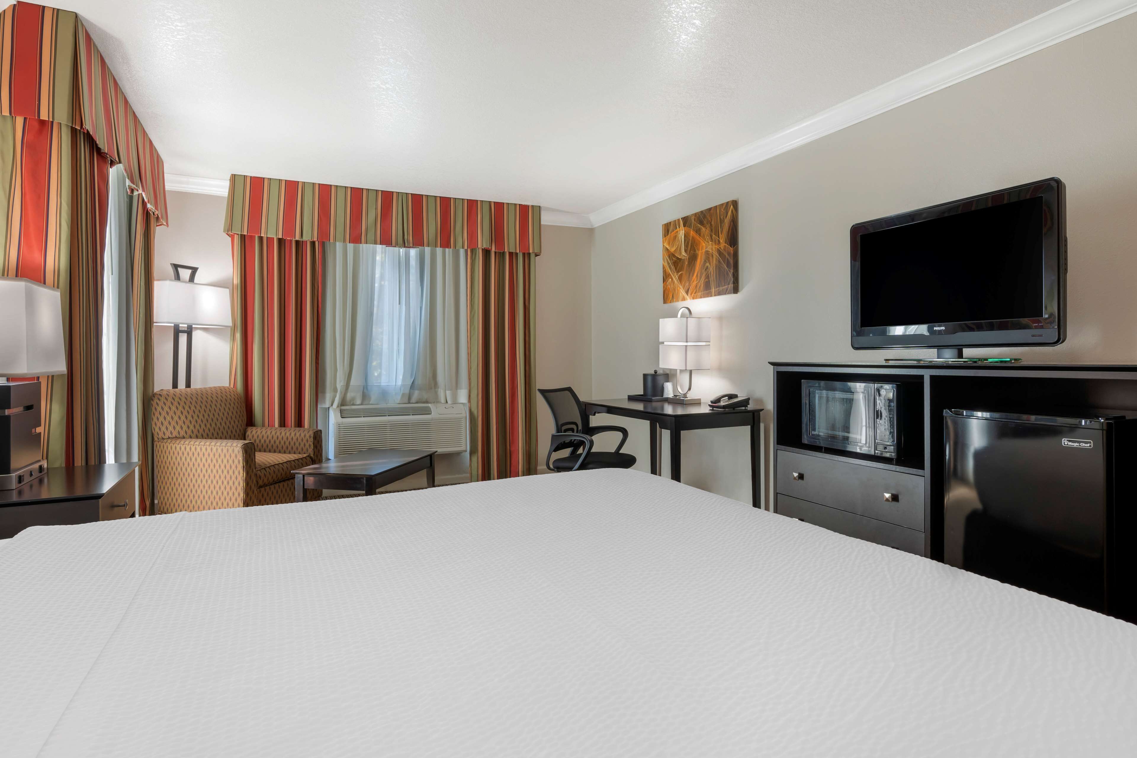 A hotel room with a large bed, a desk with a lamp, a television, a refrigerator, and a chair.